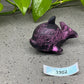 a purple fish figurine sitting on the ground next to a plant